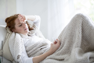 Frau mit Grippe liegt mit Fieberthermometer in der Hand auf dem Sofa.